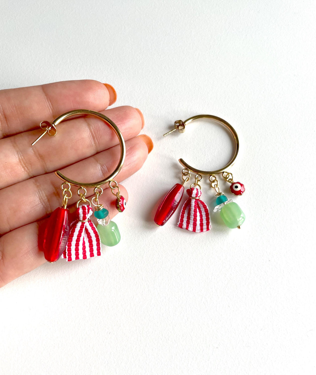 A pair of gold hoop earrings with dangling red and white striped tassels, red beads, and light green beads, shown on a person's hand against a plain white background.