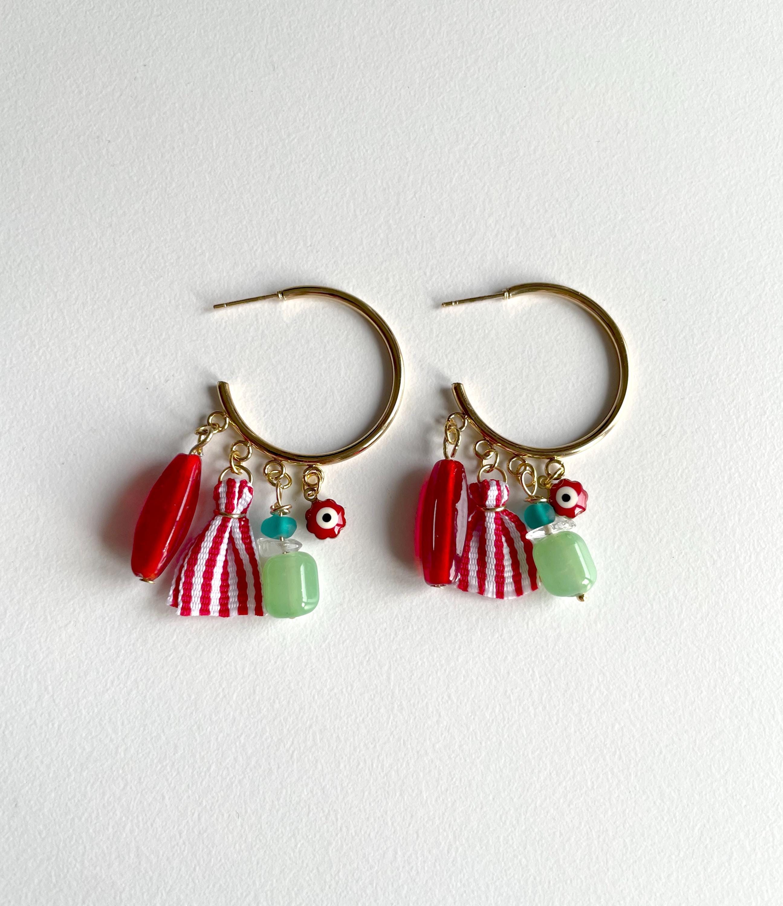 A pair of gold hoop earrings with dangling charms, including red beads, a red-and-white striped tassel, a light green bead, and small floral charms, displayed on a textured white background.