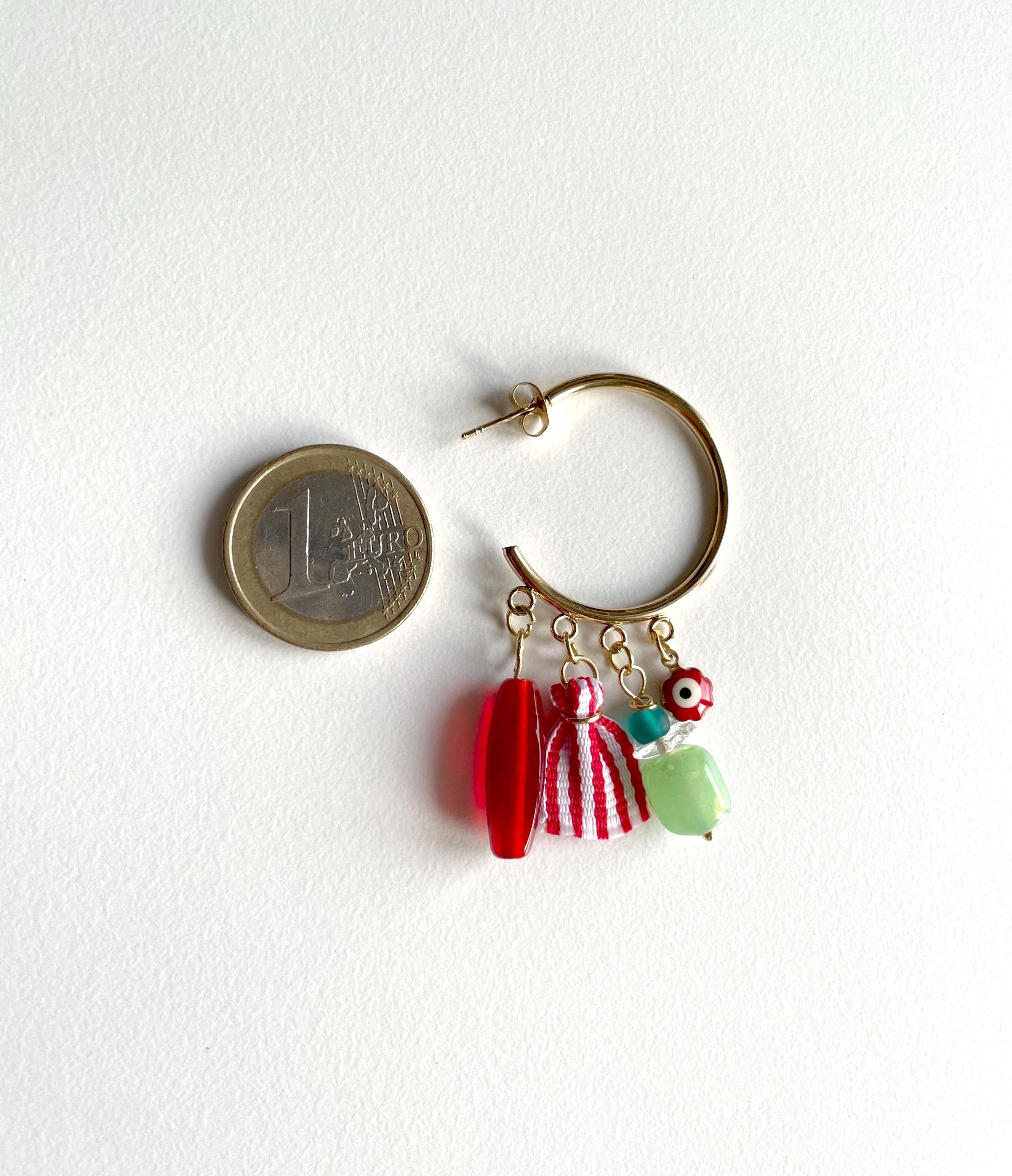 A gold hoop earring with three dangling charms: a red bead, a red-and-white striped bead, and a light green bead, shown next to a 1 Euro coin for scale on a white background.