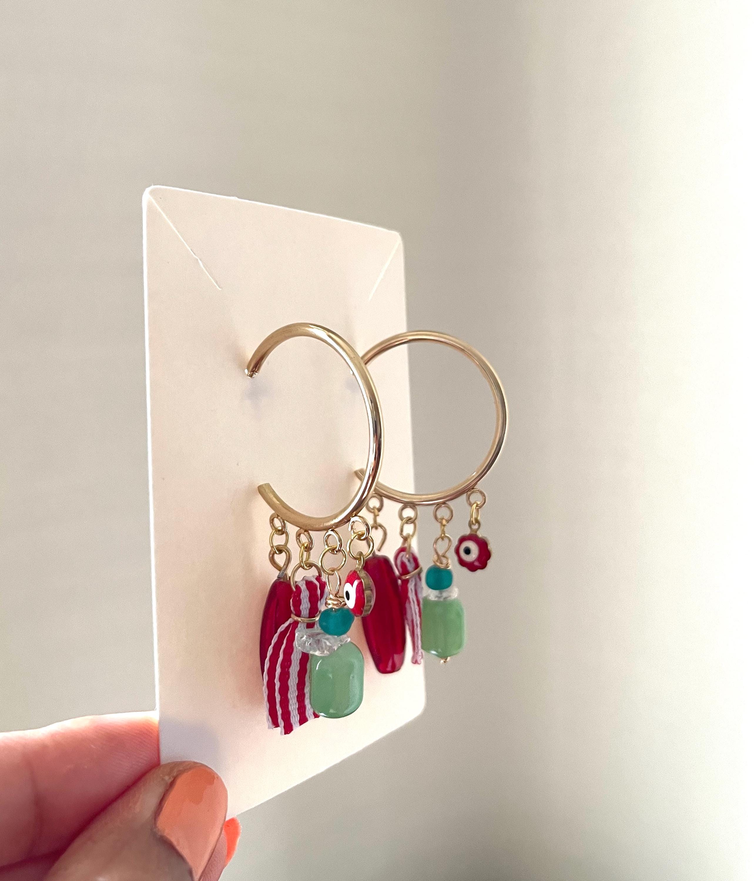 Gold hoop earrings with dangling red and green evil eye charms, displayed on a white card held by a hand.