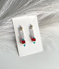 A pair of clear cylindrical drop earrings with small red cube beads, turquoise heart beads, and gold posts, displayed on a white card over a white faux fur background.