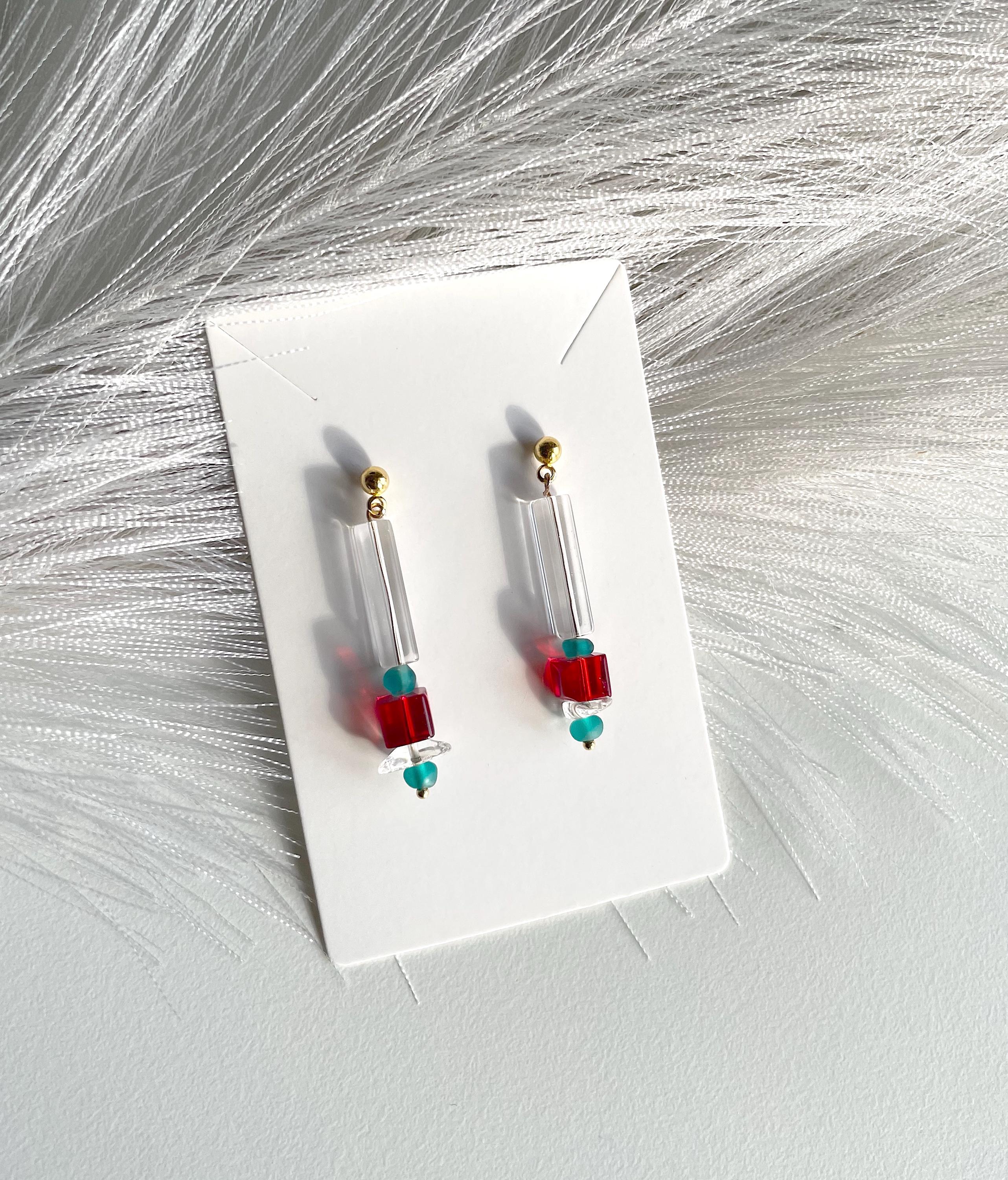 A pair of clear cylindrical drop earrings with small red cube beads, turquoise heart beads, and gold posts, displayed on a white card over a white faux fur background.
