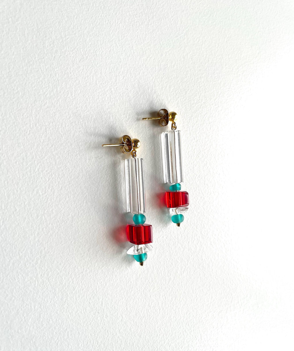 A pair of drop earrings with clear cylindrical beads, red square beads, and small turquoise round beads, hanging against a white textured wall.
