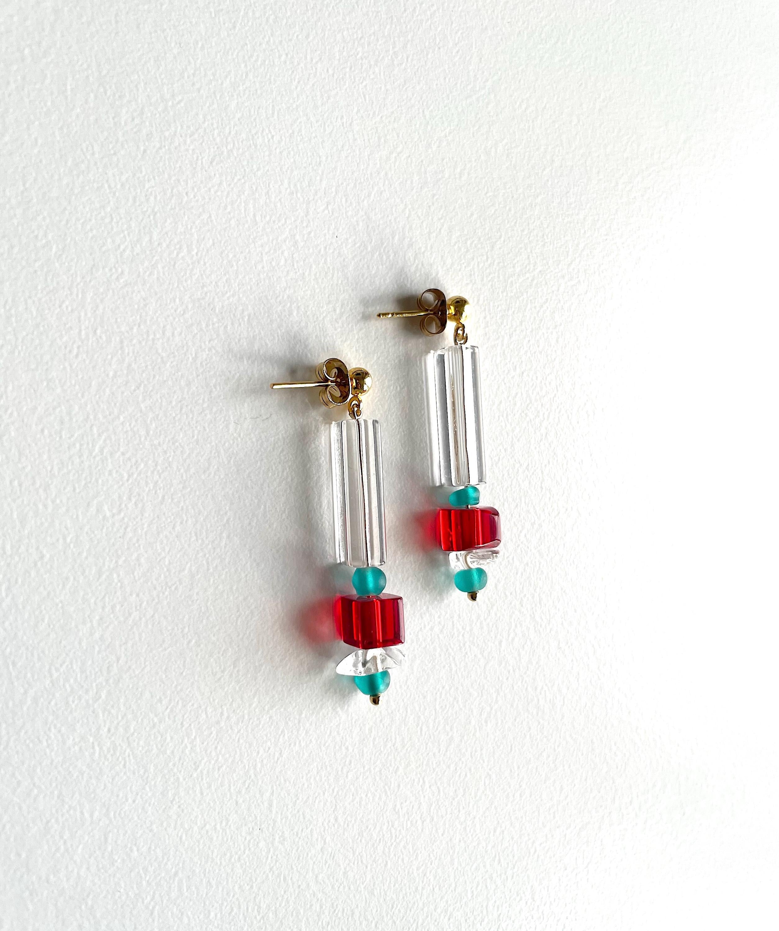 A pair of drop earrings with clear cylindrical beads, red square beads, and small turquoise round beads, hanging against a white textured wall.