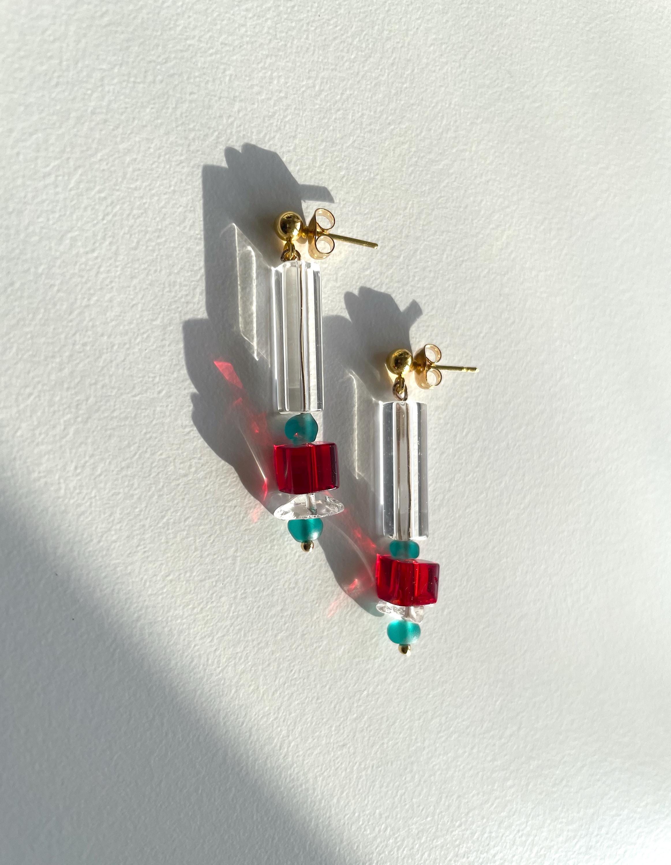 A pair of drop earrings with clear acrylic tubes, red square beads, and small teal beads, hanging on gold posts against a white wall with strong sunlight and shadows.