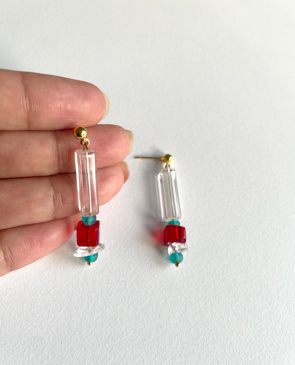 A pair of handmade dangle earrings held by a hand against a plain white background. The earrings feature clear rectangular acrylic beads, a red square bead, a small turquoise bead, and gold-tone posts.