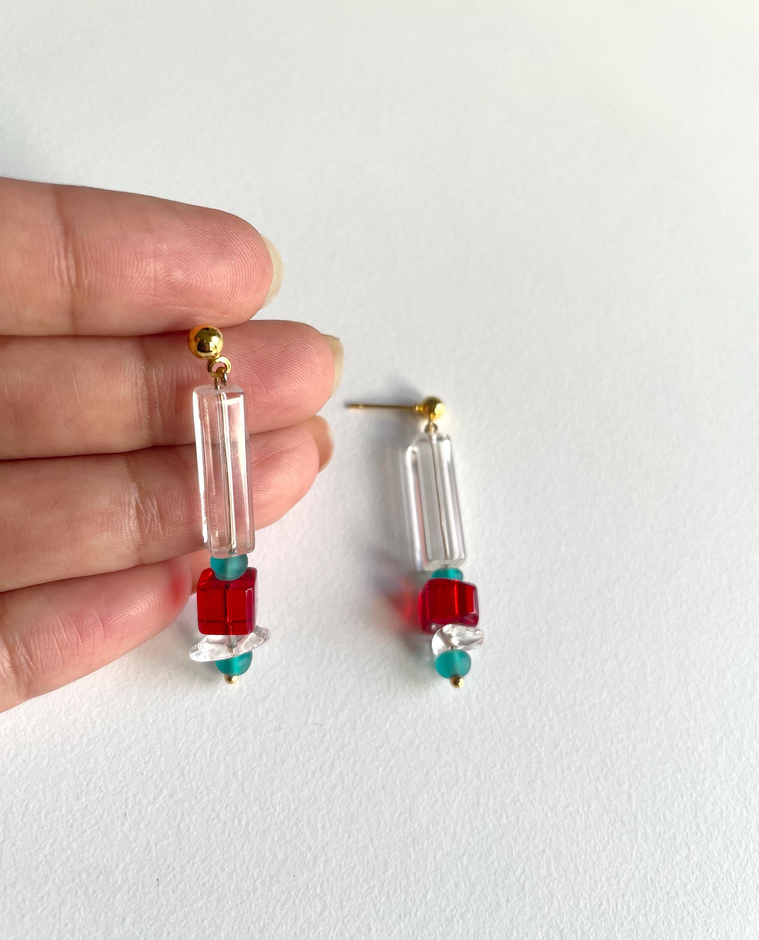 A pair of handmade dangle earrings held by a hand against a plain white background. The earrings feature clear rectangular acrylic beads, a red square bead, a small turquoise bead, and gold-tone posts.