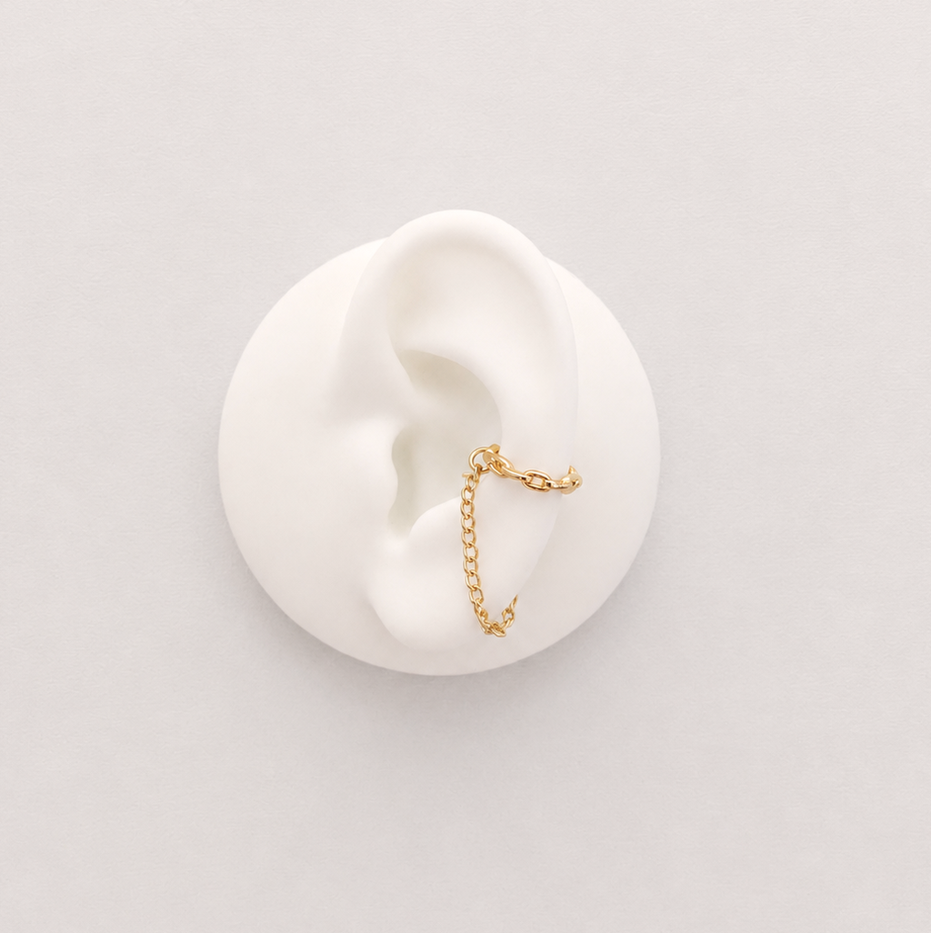A delicate rose gold ear cuff with a small dangling chain charm, displayed on a white ear-shaped mannequin against a light gray background.