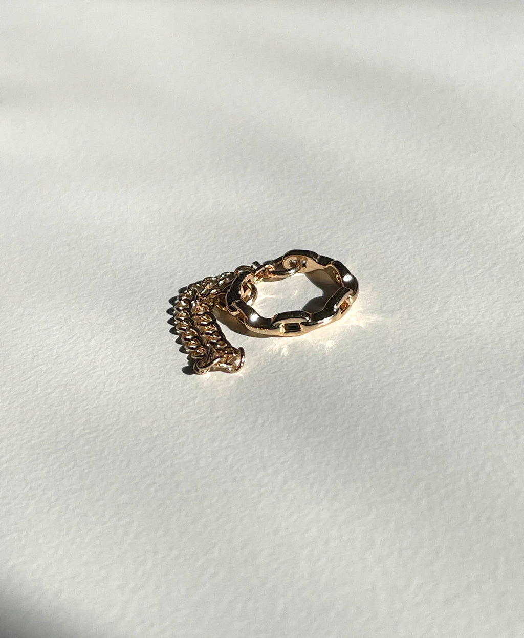 A rose gold cuff earring, wavy outer band and a matching curb chain extension, photographed on a textured off-white surface with strong natural shadows.