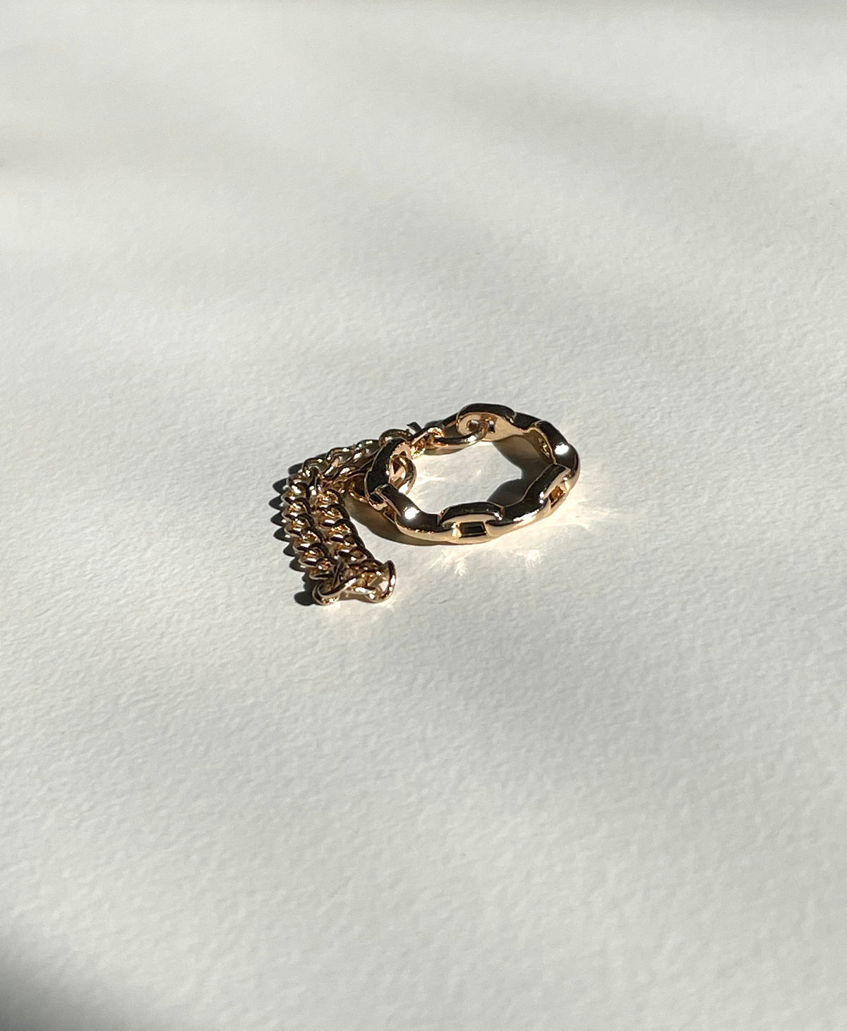 A rose gold cuff earring, wavy outer band and a matching curb chain extension, photographed on a textured off-white surface with strong natural shadows.