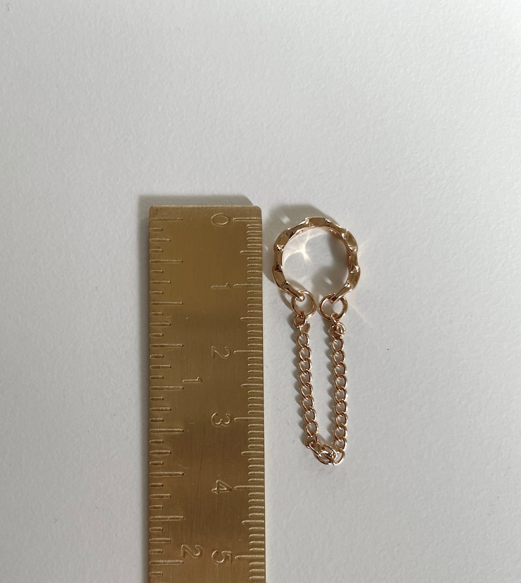A small rose gold cuff earring with a short chain extension, shown next to a brass ruler for scale on a plain white background.