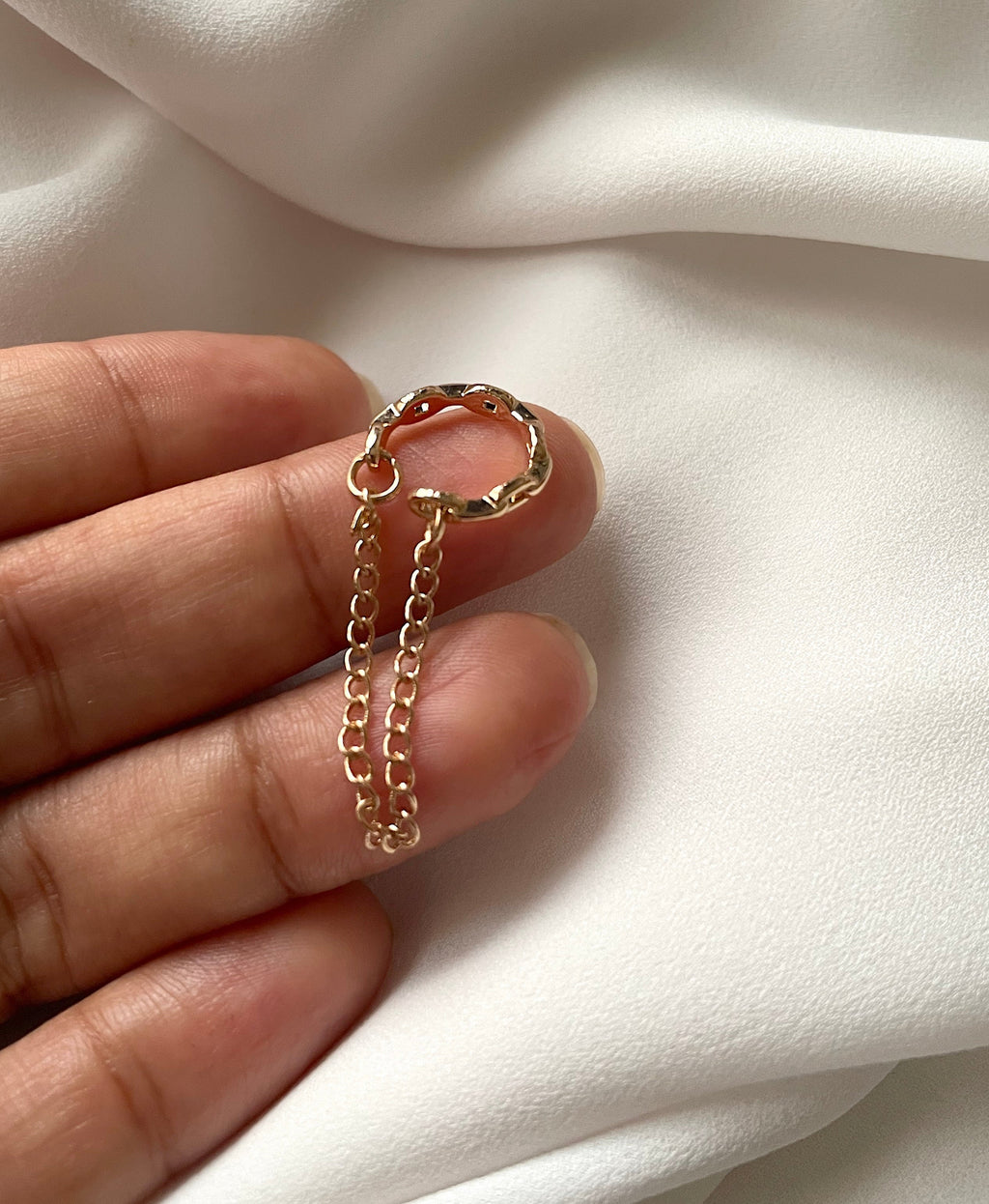 A gold cuff earring with a textured circular top and a short dangling chain, held between fingers against a soft white fabric background.
