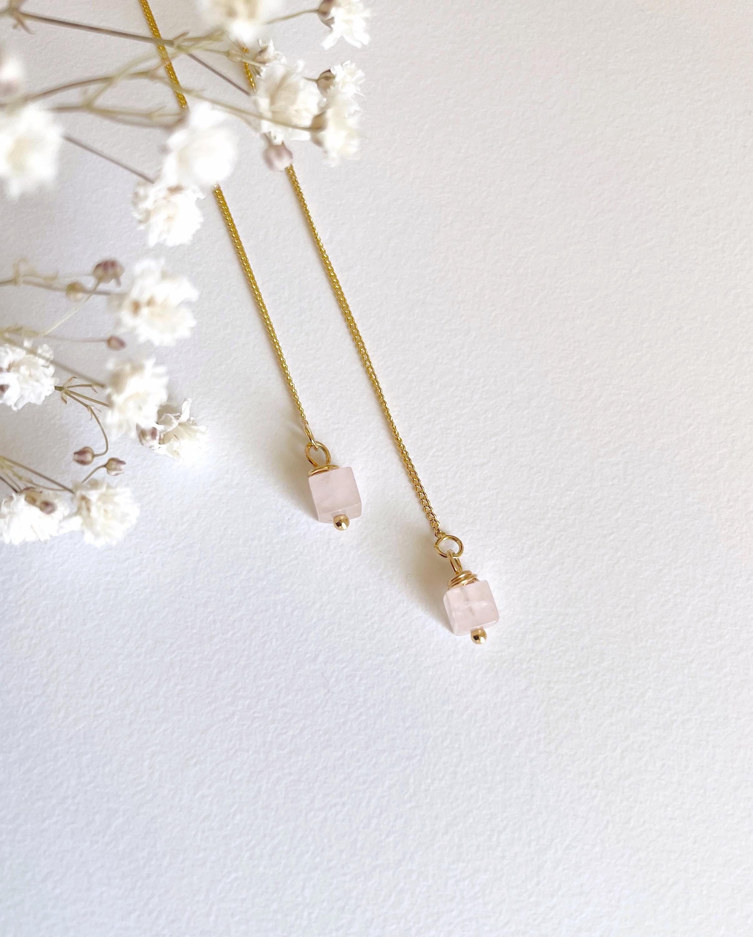 A pair of delicate gold chain necklaces with small rectangular pale pink stone pendants, styled with white baby's breath flowers on a textured white background.