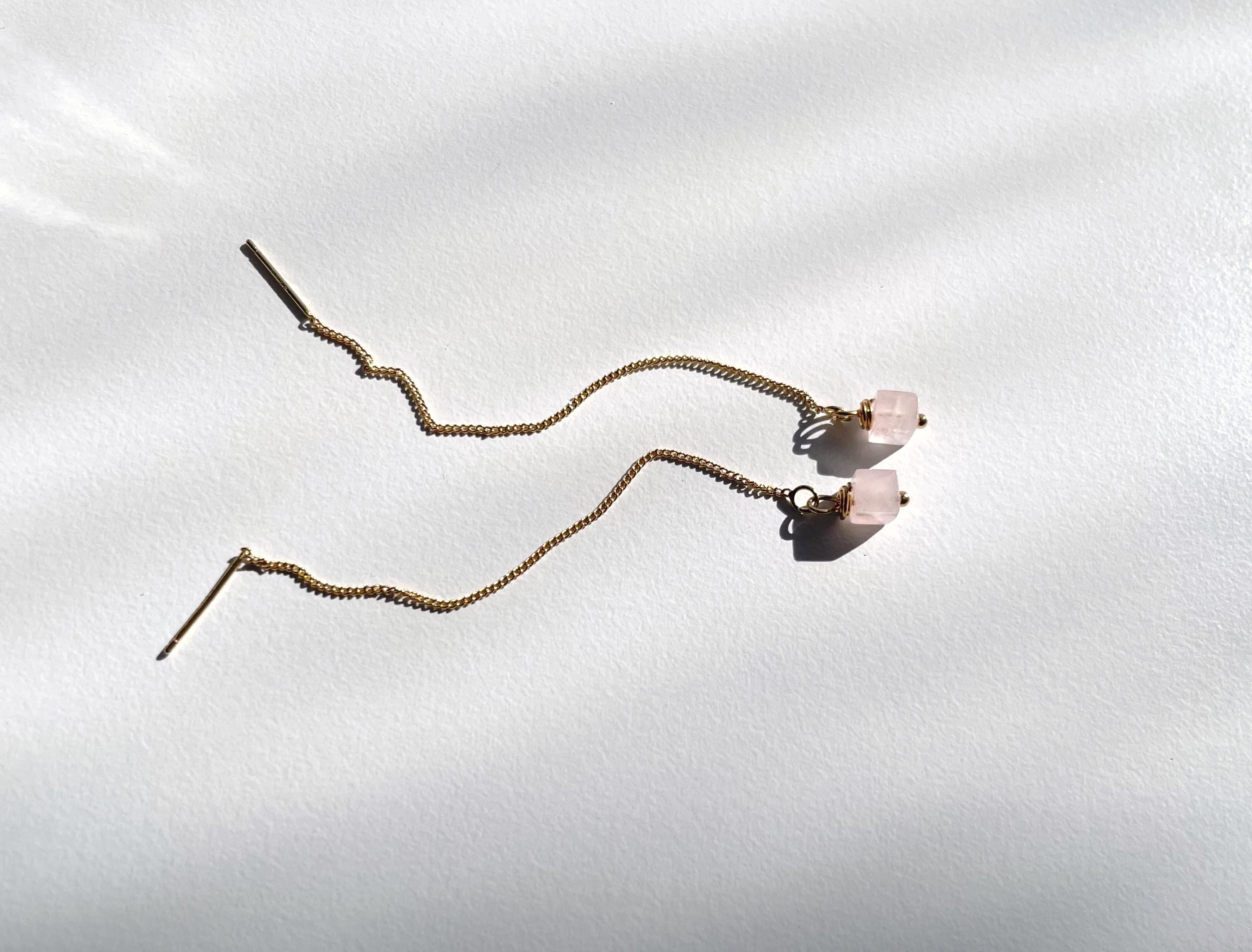 A pair of long, wavy gold-toned chain earrings with small pale pink bead accents, displayed on a white textured background with soft shadows.