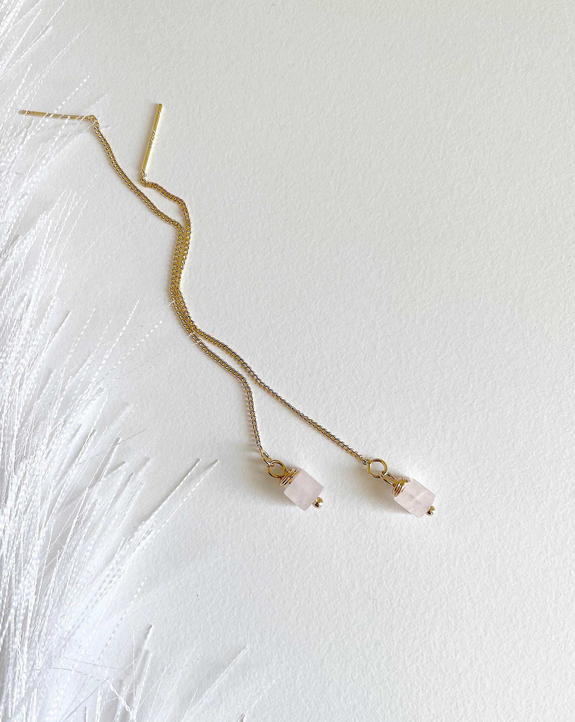 A delicate gold chain necklace with two small dangling pink stone beads, laid flat on a textured white surface with white feather-like decor on the left.
