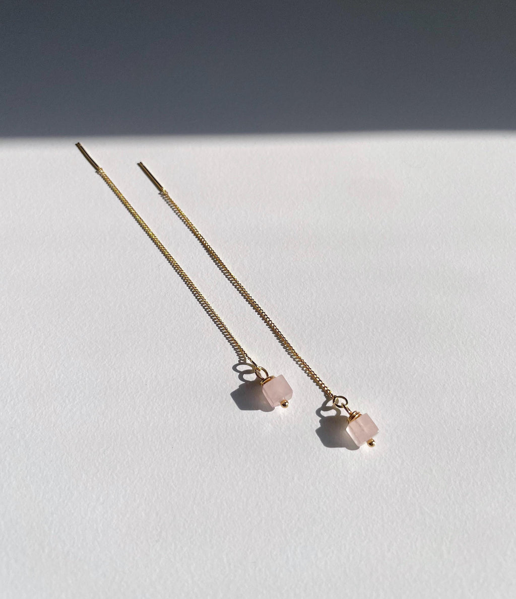 A pair of long gold chain threader earrings with small pale pink rose quartz square bead charms, photographed on a white surface with a dark shadow across the upper background.