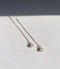 A pair of long gold chain threader earrings with small pale pink rose quartz square bead charms, photographed on a white surface with a dark shadow across the upper background.