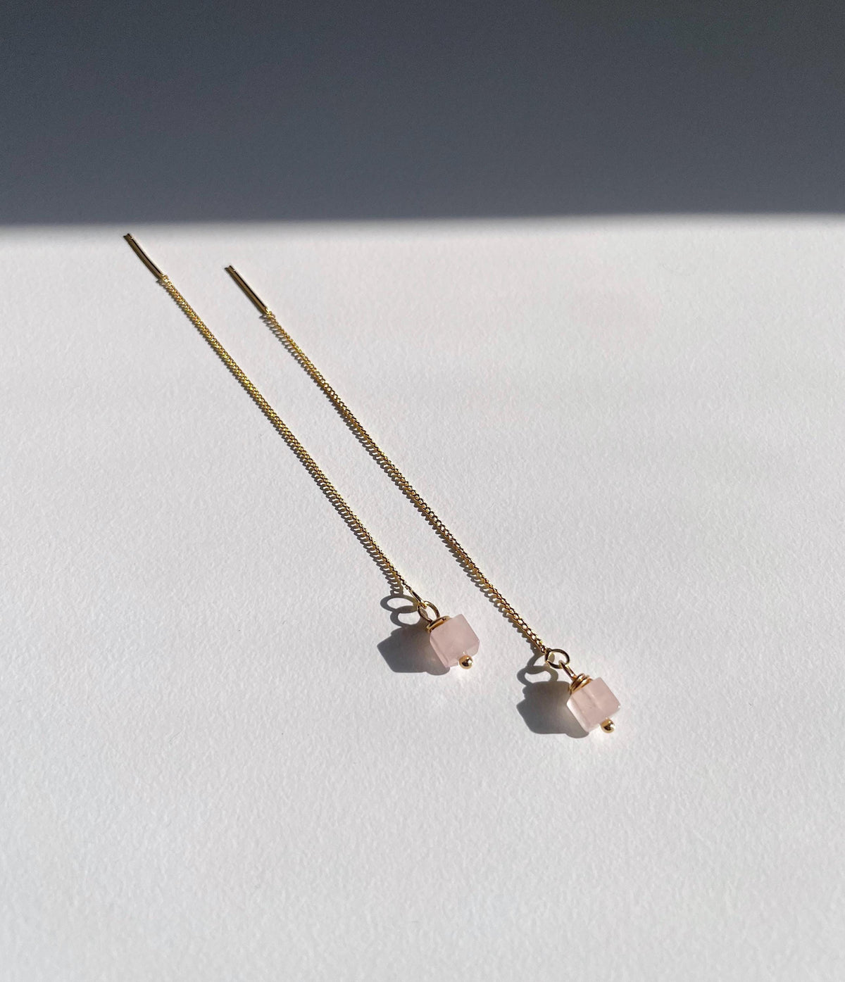 A pair of long gold chain threader earrings with small pale pink rose quartz square bead charms, photographed on a white surface with a dark shadow across the upper background.