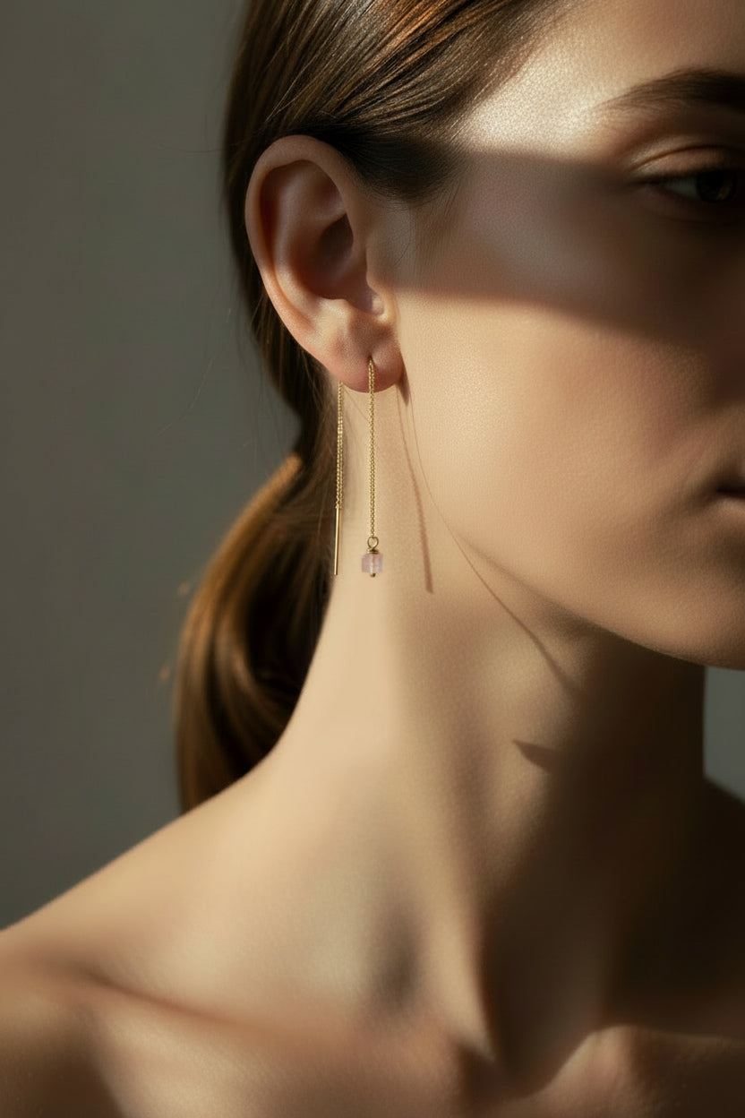 A close-up side profile of a woman wearing a delicate gold dangle threader earring with a small rose quartz square bead, set against a dark gray background with dramatic shadow play.
