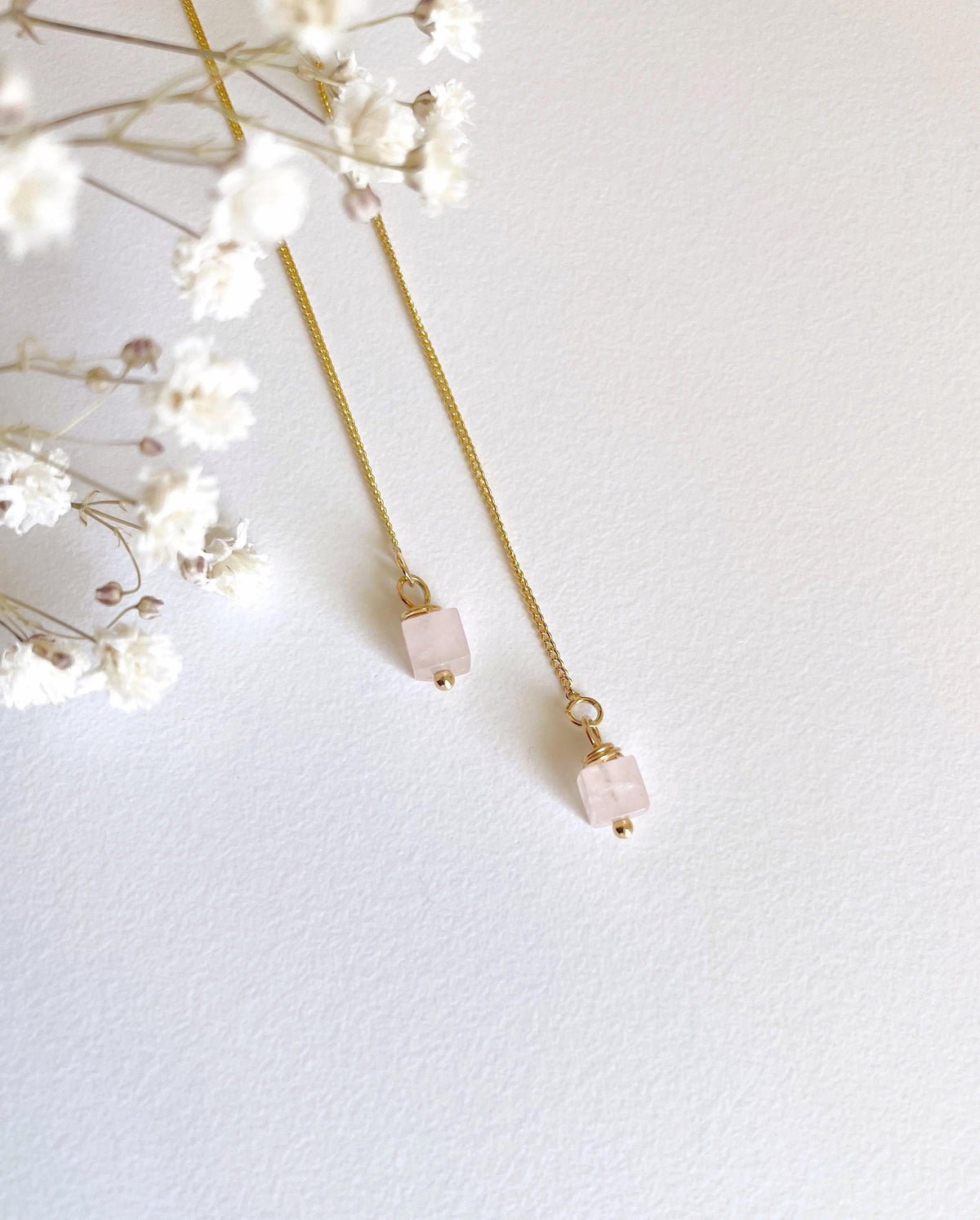A pair of delicate gold chain threader earrings with small square pale pink rose quartz natural stone beads, styled with white baby's breath flowers on a textured white background.