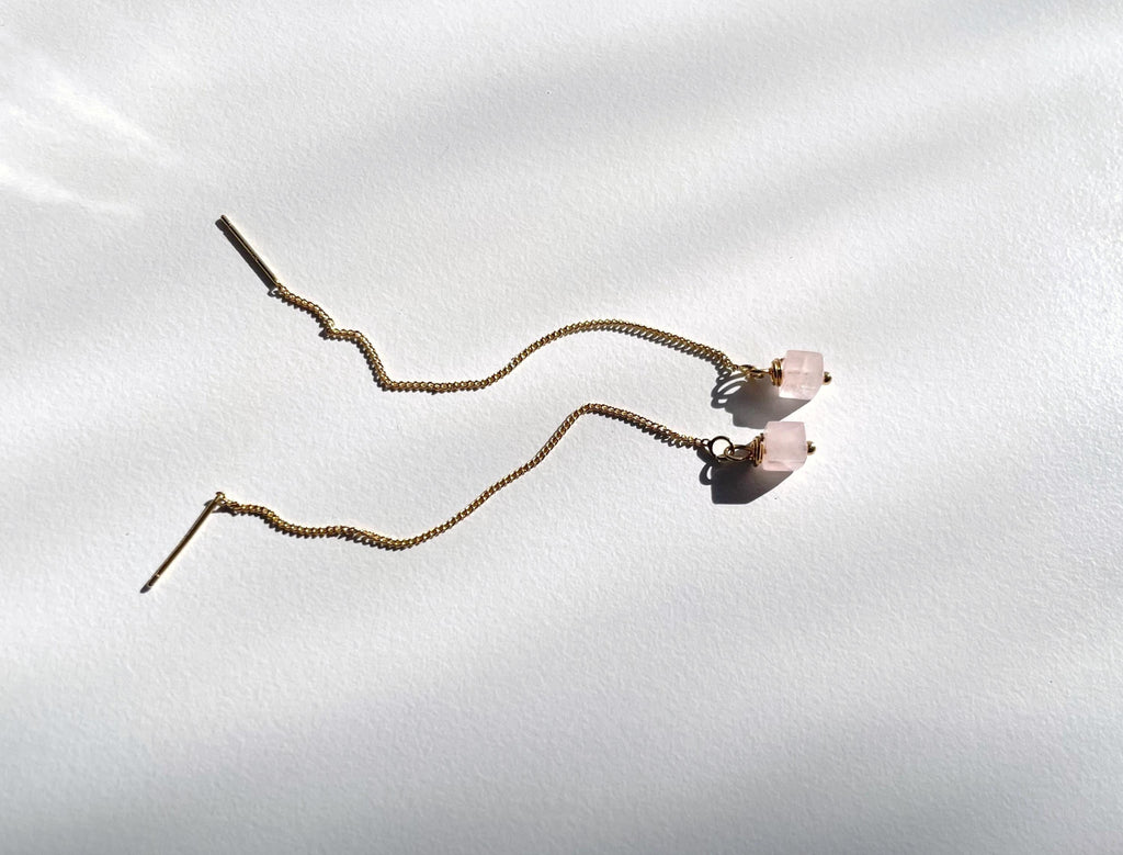 A pair of long, wavy gold plated  threader earrings with small natural rose quartz bead accents, displayed on a white textured background with soft shadows.