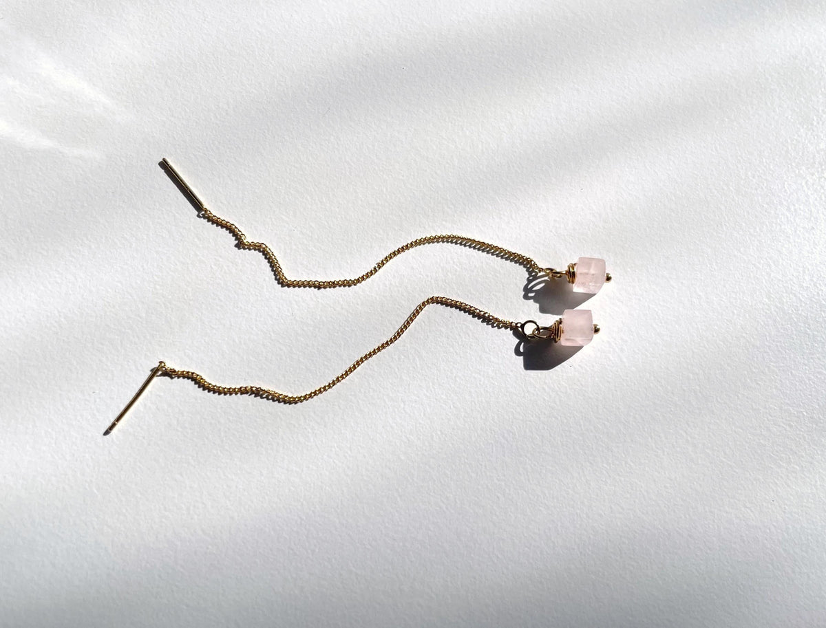 A pair of long, wavy gold plated  threader earrings with small natural rose quartz bead accents, displayed on a white textured background with soft shadows.
