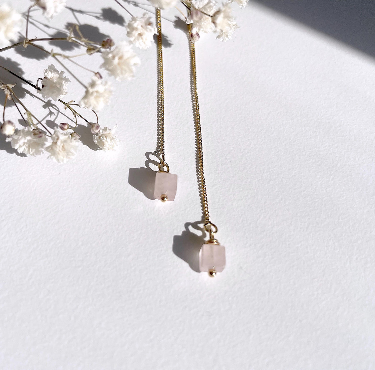 A pair of long golden threader earrings with pale pink square rose quartz natural stone pendants, styled with white baby's breath flowers on a textured white surface under bright sunlight.