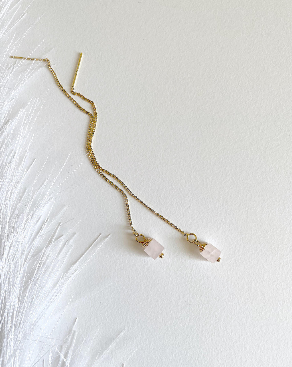 A pair of delicate golden chain earrings with small dangling light pink square stone beads, laid flat on a textured white surface with white feather-like decor on the left.
