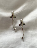 Two polished silver dolphin-shaped shark earrings displayed on a textured white fabric background.
