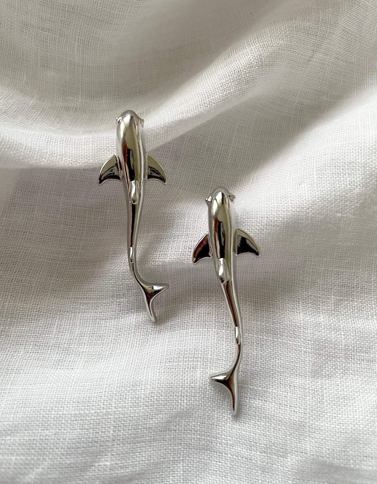 Two polished silver dolphin-shaped shark earrings displayed on a textured white fabric background.