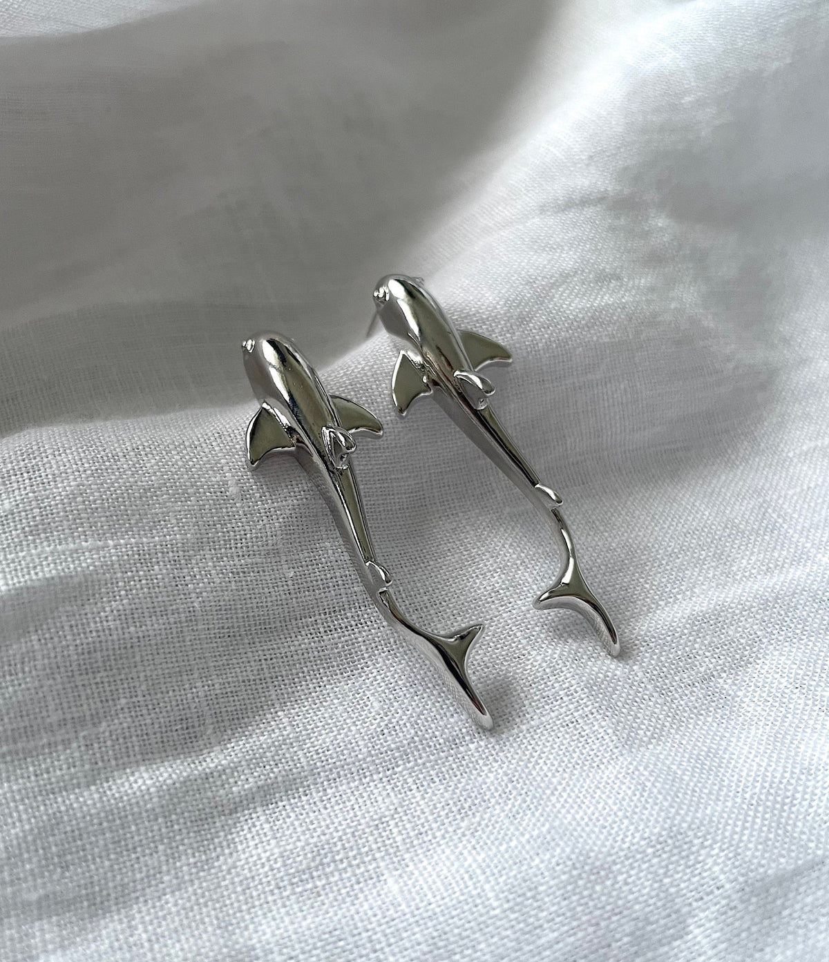A pair of silver shark-shaped earrings resting on a textured white fabric background.