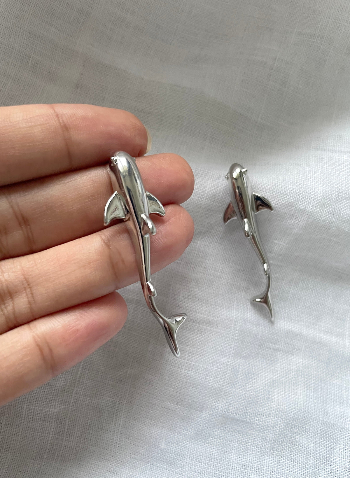 Two small polished silver shark-shaped earrings, shown with a person's fingers for scale against a light fabric background.