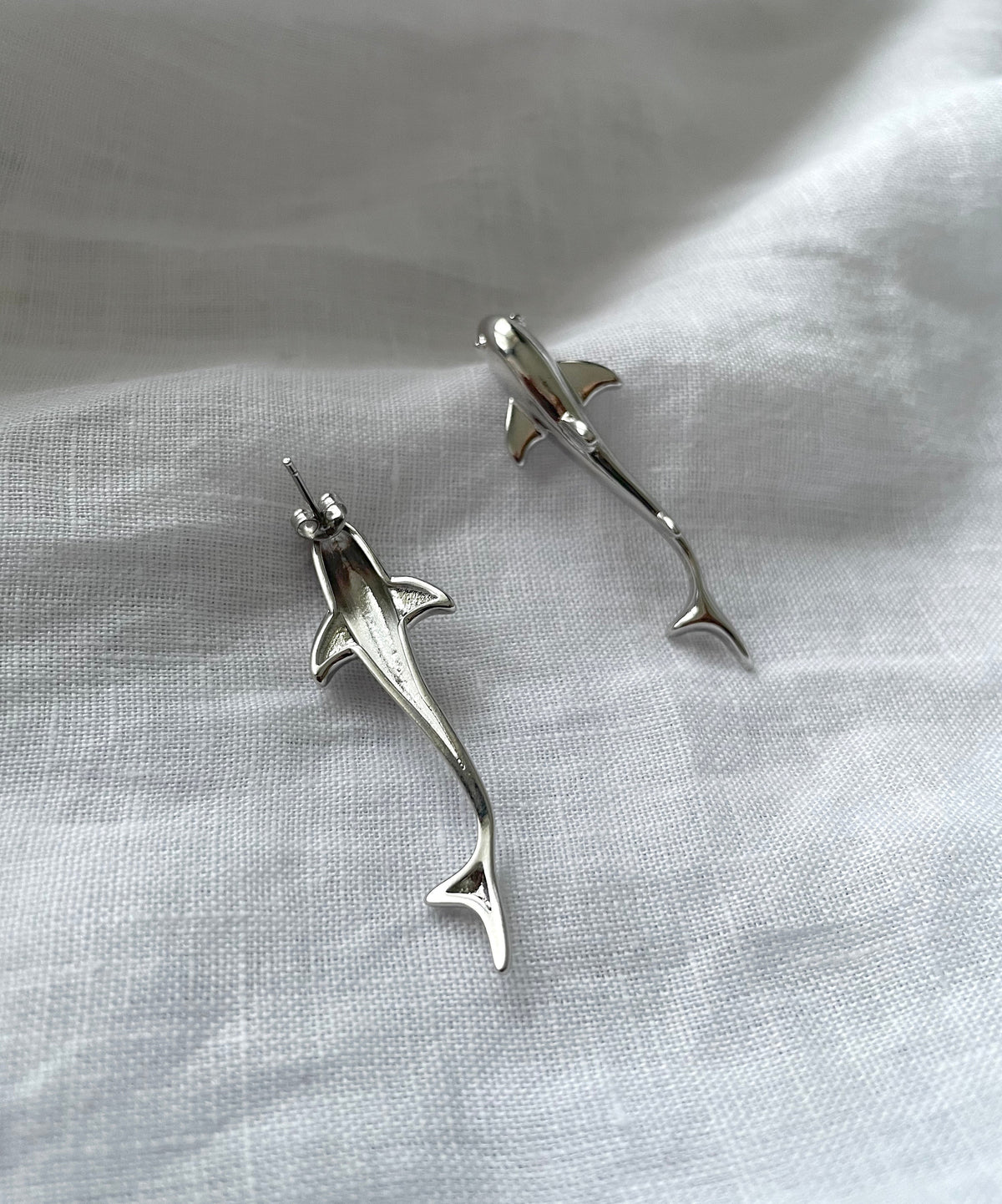 A pair of silver-toned shark-shaped stud earrings resting on a textured white fabric background.