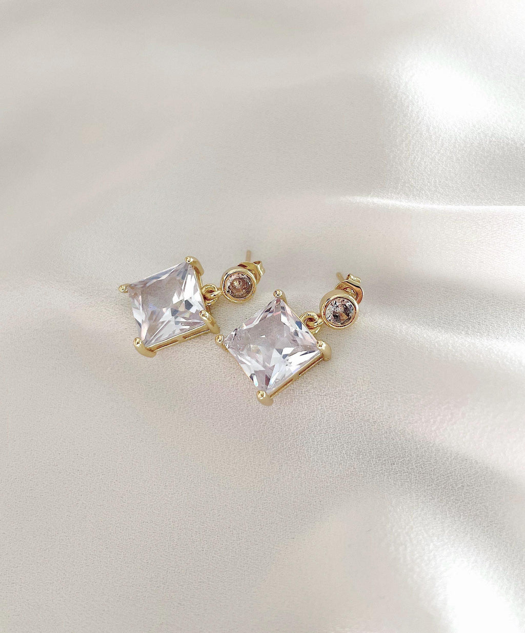 A pair of gold-plated stud earrings featuring large clear square-cut crystals and smaller round clear stones, displayed on a soft, light-colored satin fabric background.