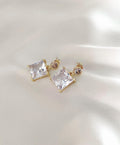 A pair of gold-plated stud earrings featuring large clear square-cut crystals and smaller round clear stones, displayed on a soft, light-colored satin fabric background.