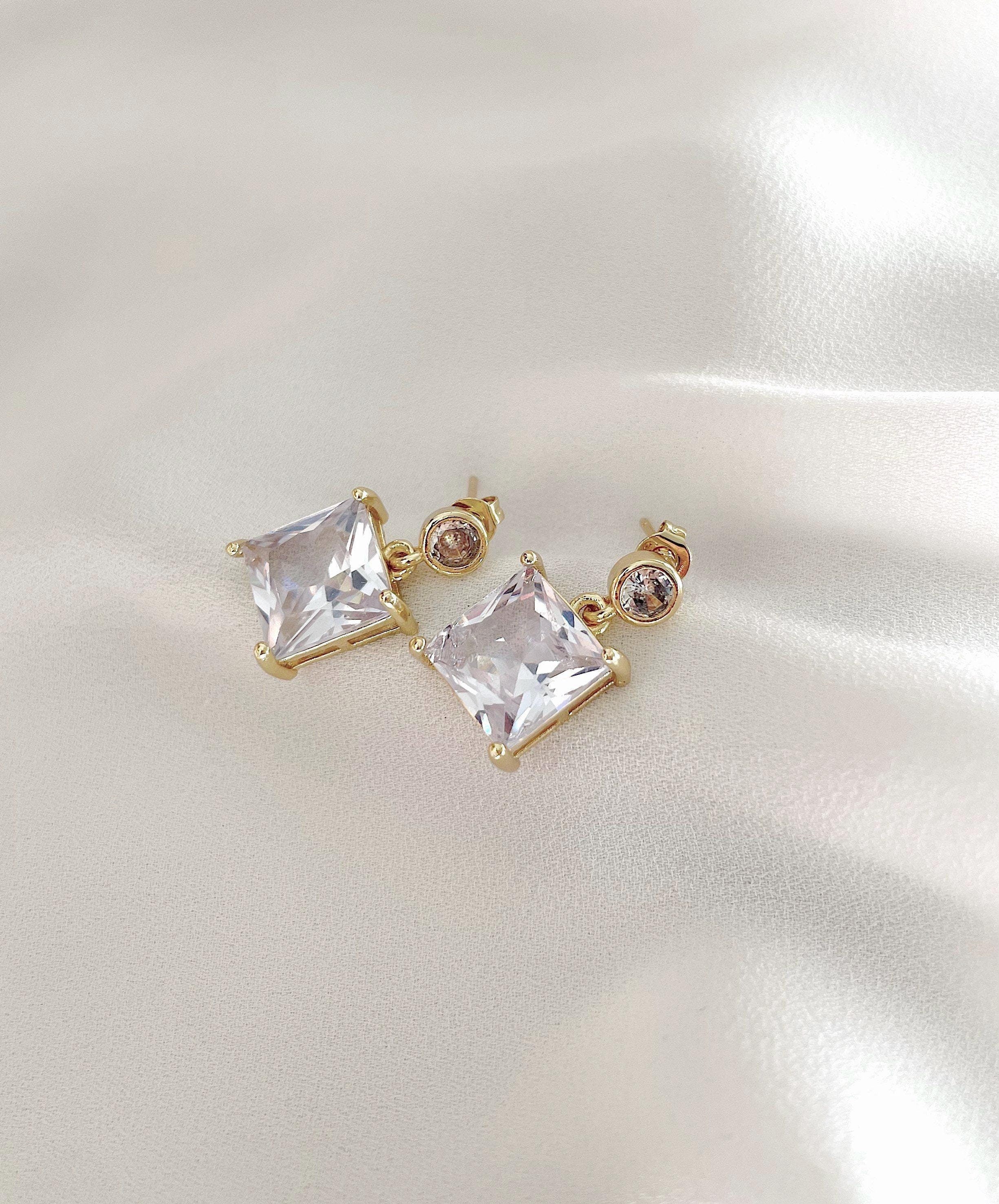 A pair of gold-plated stud earrings featuring large clear square-cut crystals and smaller round clear stones, displayed on a soft, light-colored satin fabric background.