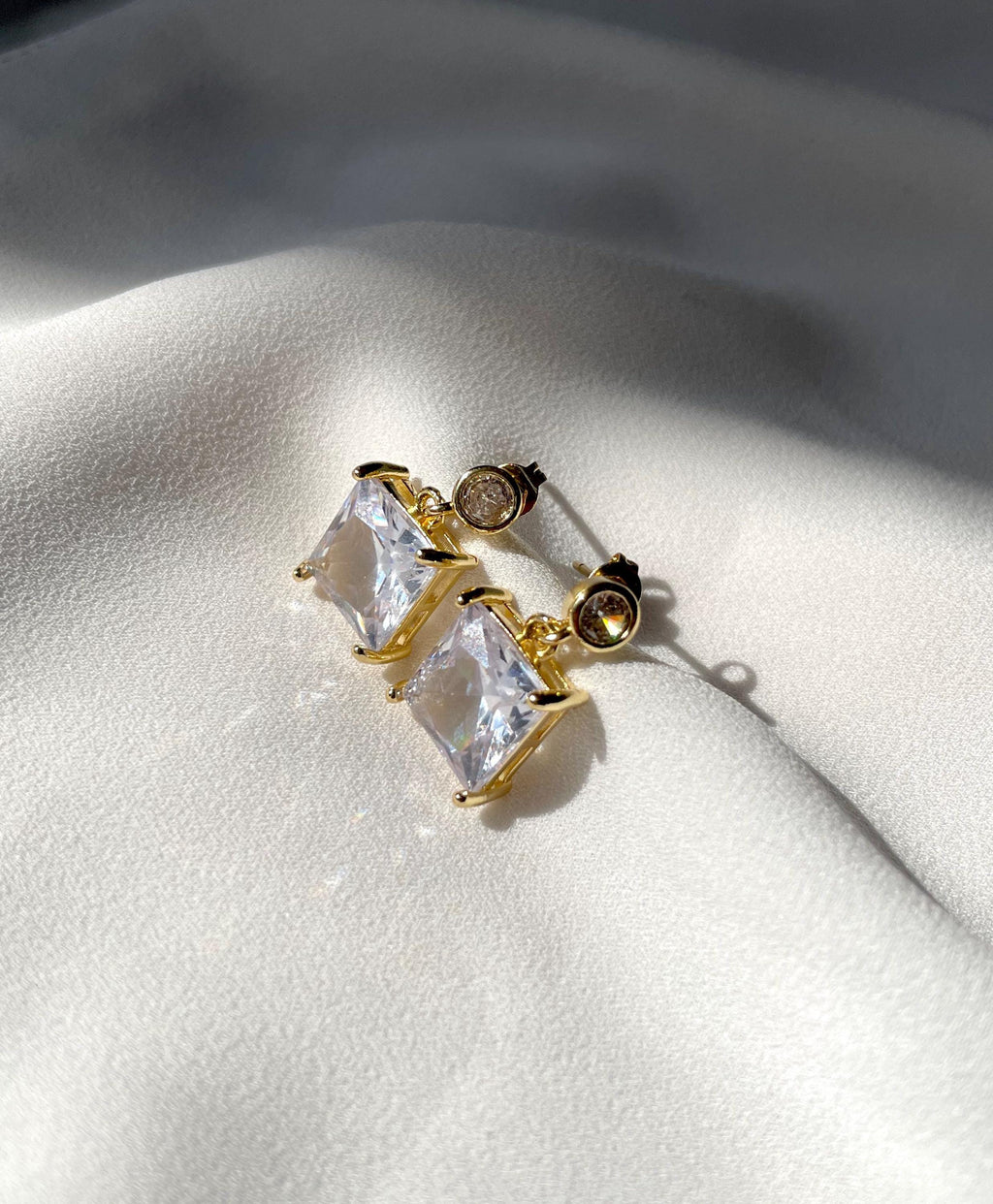 A pair of gold-toned stud earrings with large clear rectangular stones and a small round clear stone, displayed on a white textured fabric background.