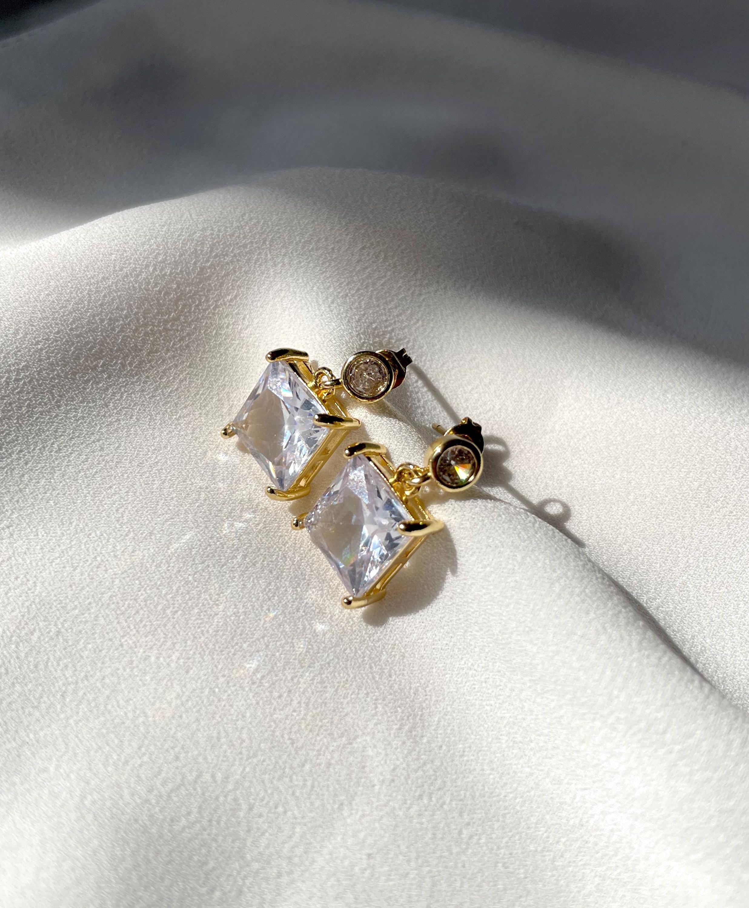 A pair of gold-toned stud earrings with large clear rectangular stones and a small round clear stone, displayed on a white textured fabric background.