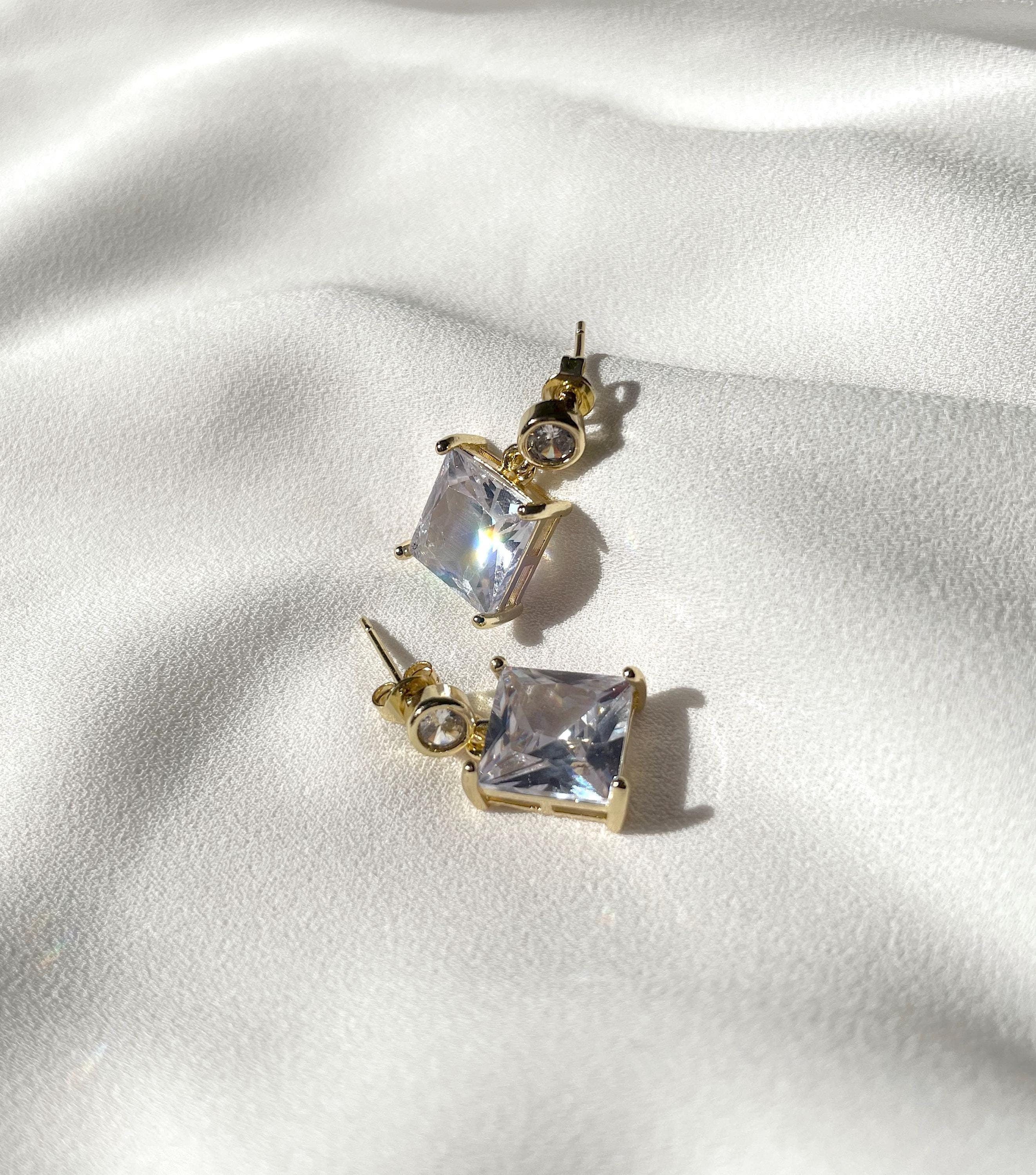 A pair of gold stud earrings with square clear gemstones, displayed on a textured white fabric background.