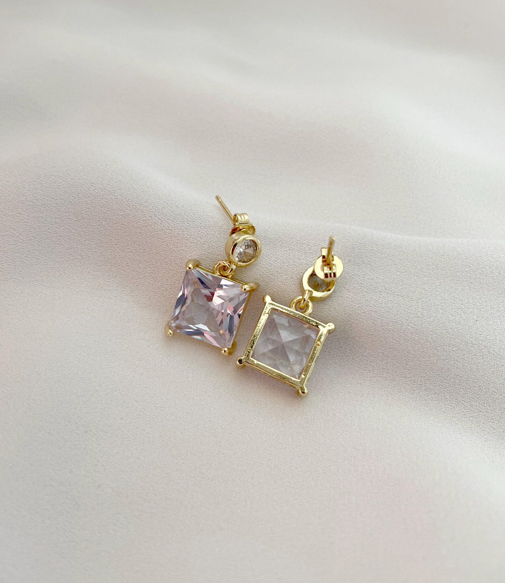 A pair of gold stud earrings with square-cut pale pink and clear stones, displayed on a soft, light-colored fabric background.