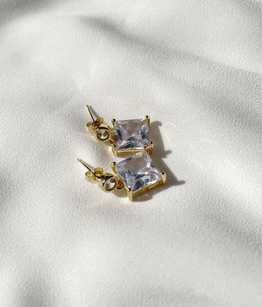 A pair of gold stud earrings with square clear gemstones, resting on a textured white fabric background.