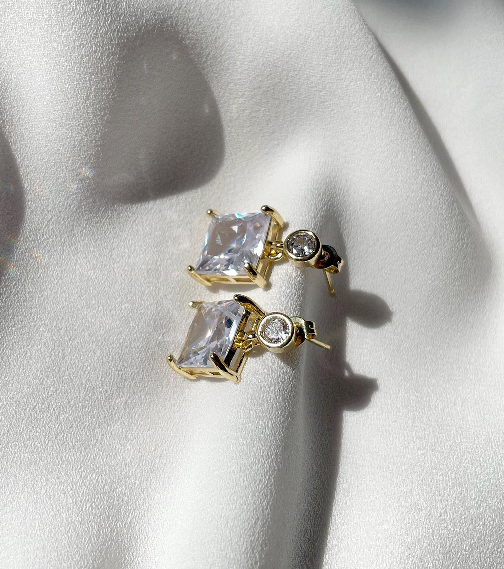 A pair of gold-toned stud earrings with rectangular clear gemstones and a small round clear stone, displayed on a white fabric surface.