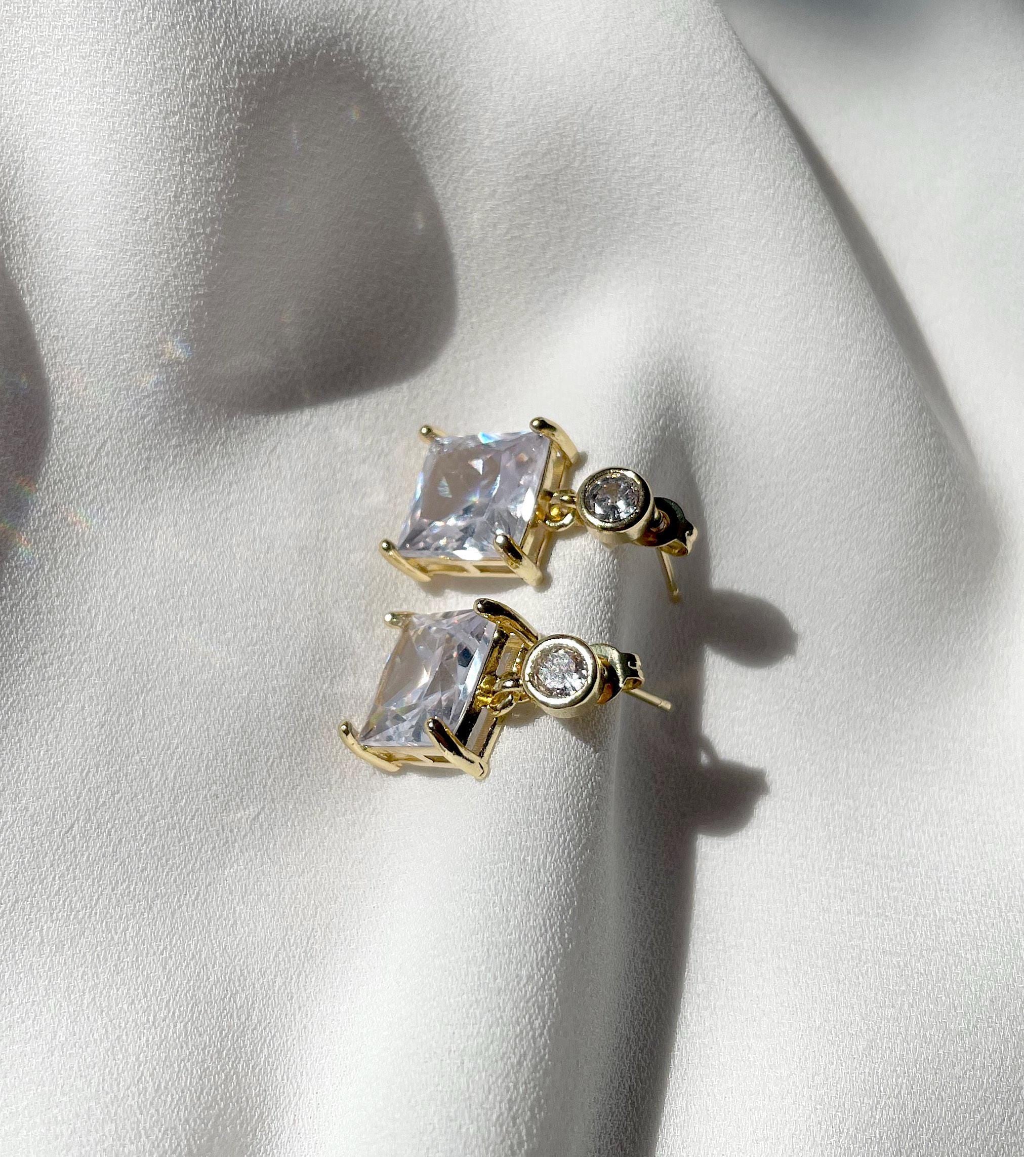 A pair of gold-toned stud earrings with rectangular clear gemstones and a small round clear stone, displayed on a white fabric surface.