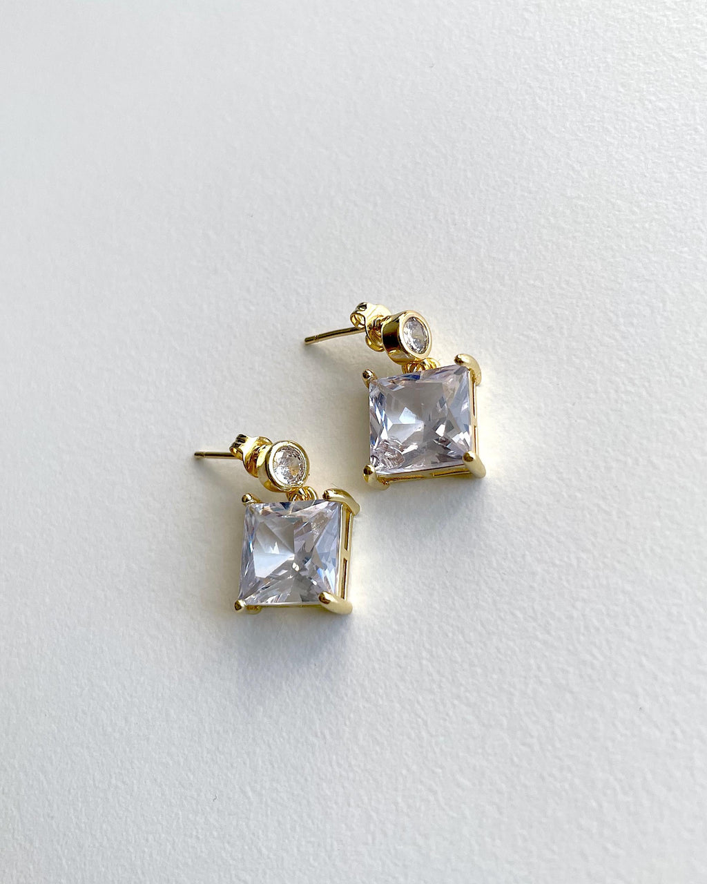 A pair of gold-plated stud earrings featuring a square clear crystal pendant and a small round clear crystal accent, displayed on a textured white surface.