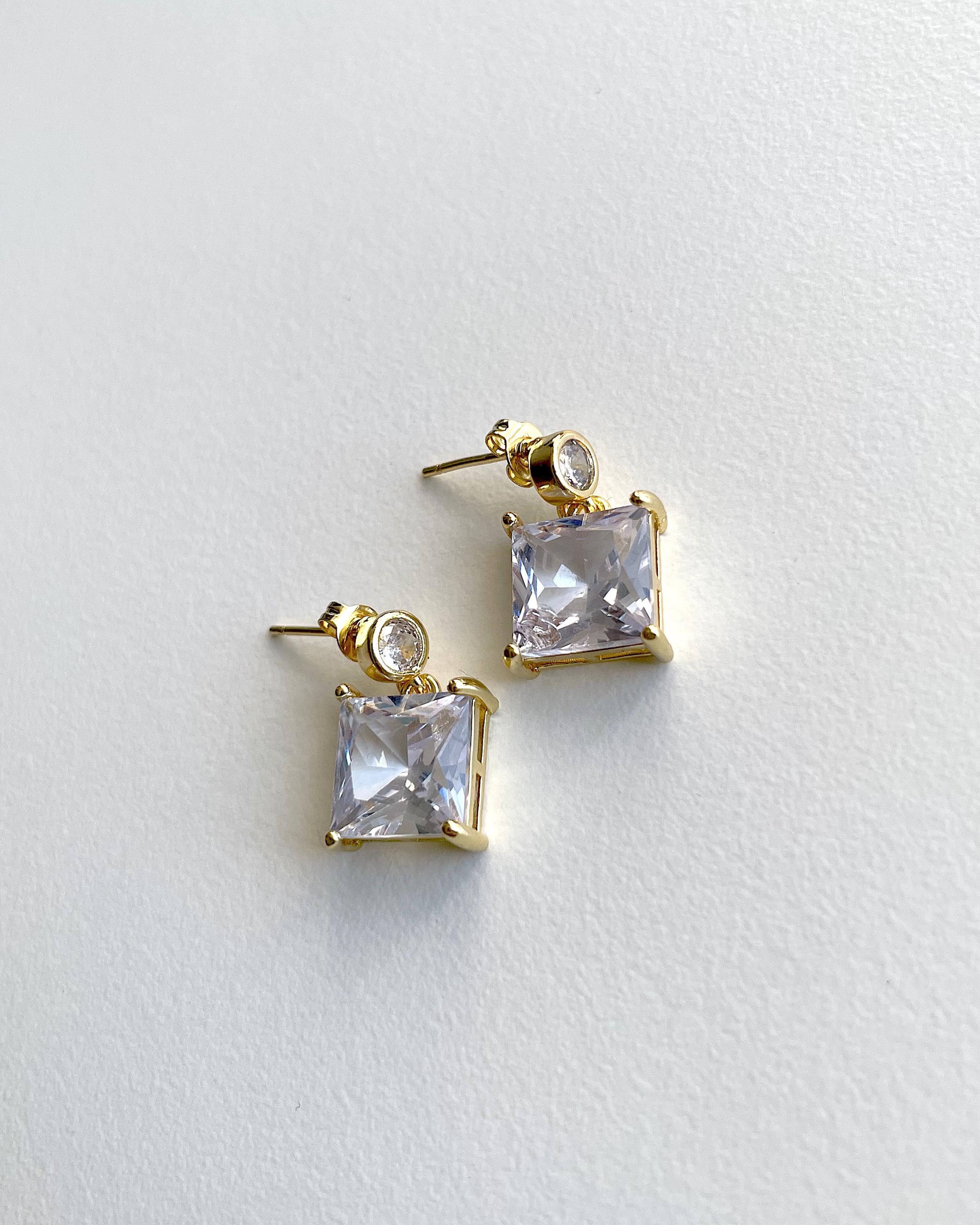 A pair of gold-plated stud earrings featuring a square clear crystal pendant and a small round clear crystal accent, displayed on a textured white surface.