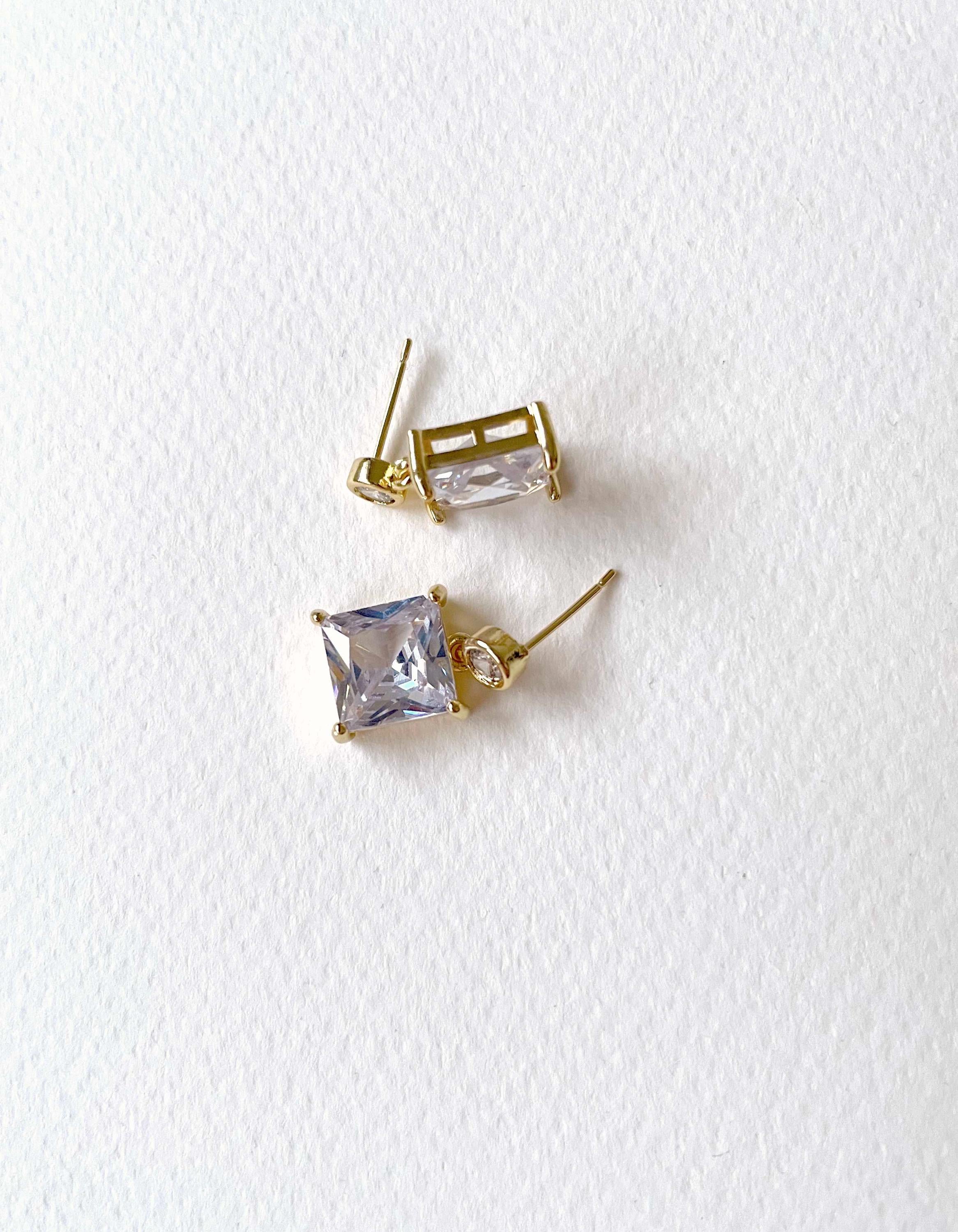 A pair of gold stud earrings featuring clear rectangular and square-cut rhinestones, displayed on a textured white background.