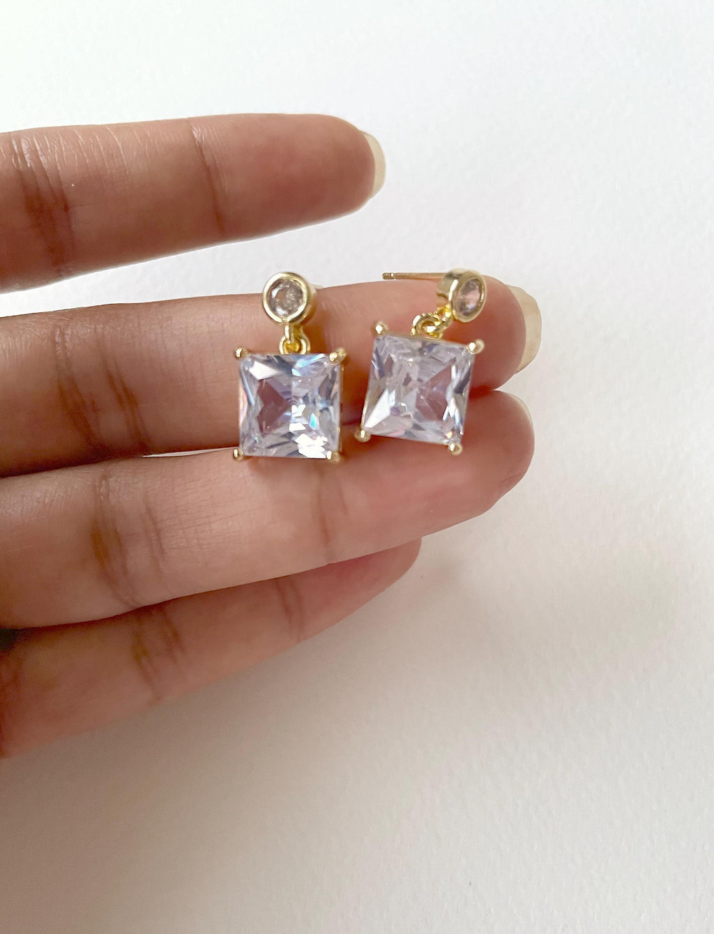 A pair of gold-plated stud earrings with square clear crystals, held on a person's fingers against a plain white background.