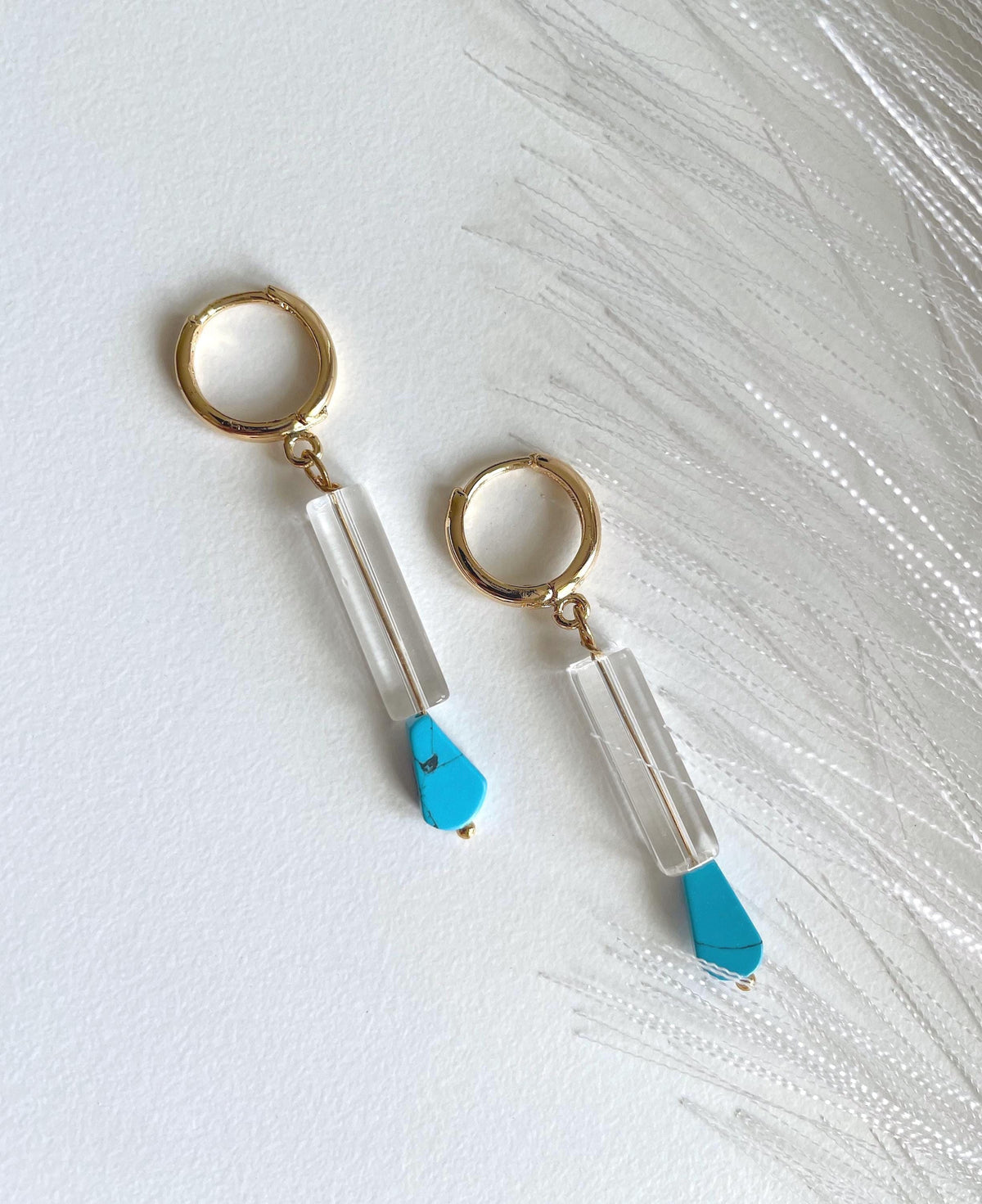 A pair of gold plated hoop drop earrings with clear cylindrical beads and bright turquoise blue teardrop pendants, displayed on a textured white surface with white feather-like strands.