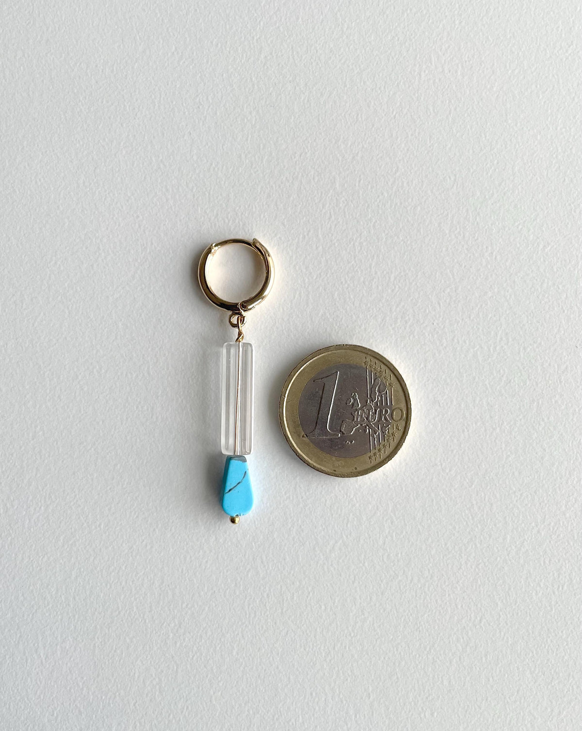 A hoop earring with a clear glass tube, and a small turquoise blue bead, shown next to a 1 Euro coin for scale on a light gray background.
