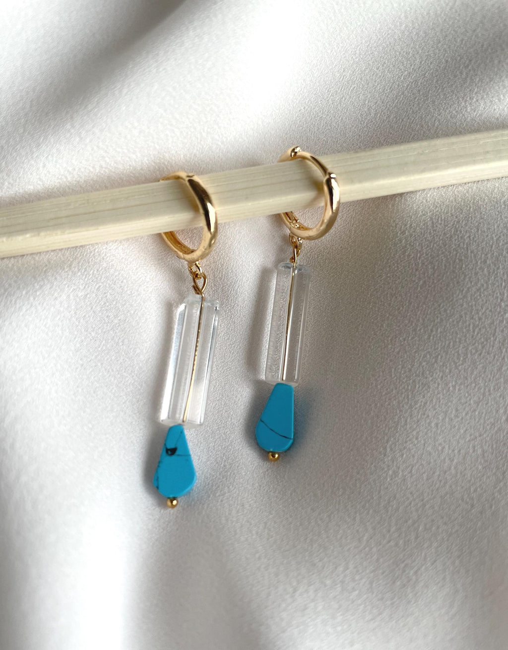A pair of gold hoop earrings with clear rectangular acrylic drops and bright turquoise blue teardrop pendants, displayed on a light wood stick against a white fabric background.